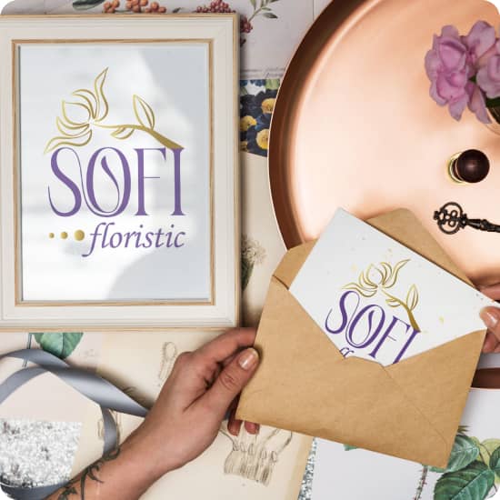 Рука тримає відкритий конверт з якого виглядає відкритка з логотипом Sofi Floristic та окремо розміщена біла рамка з роздрукованим логотипом всередині