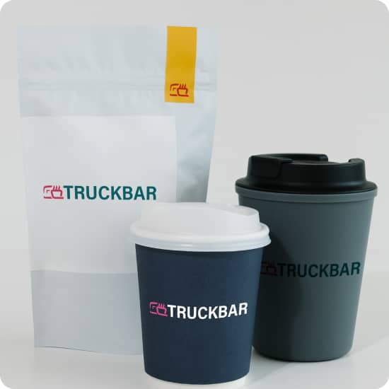 Три предмети з логотипом TRUCKBAR ресторану для водіїв: пластиковий стакан чорно-сірого кольору і чорний логотип; Пластиковий стакан біло-синього кольору і білий логотип; Пластикова упаковка для печива білого кольору з зелено-червоним лого